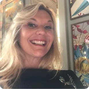Smiling blonde woman in black top with silver brooch, colourful stained-glass floral panels behind her