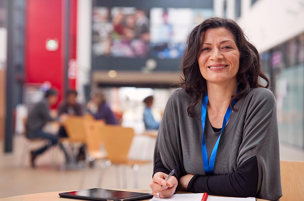 Smiling university staff member with blue lanyard at atrium table, tablet and notebook in front, students blurred behind