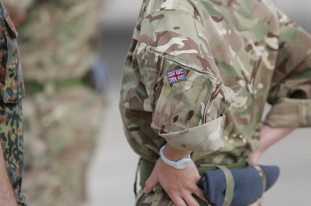 Camouflaged uniform sleeve bearing a Union Jack shoulder patch, soldier standing with hands on hips
