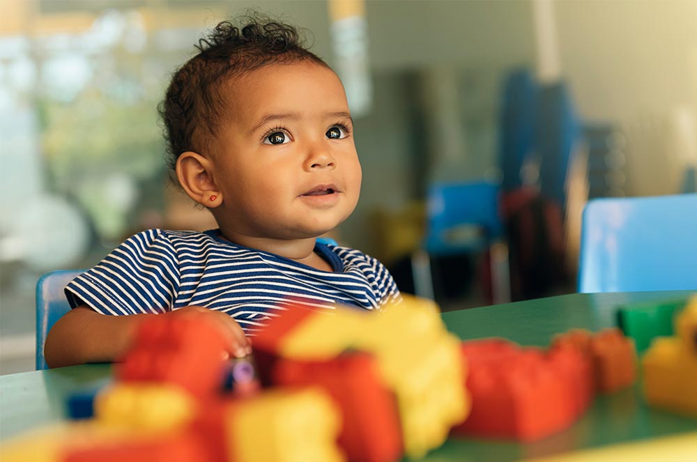 Toddler in a blue-striped top seated at a table with colourful plastic building blocks, looking up in a nursery classroom.