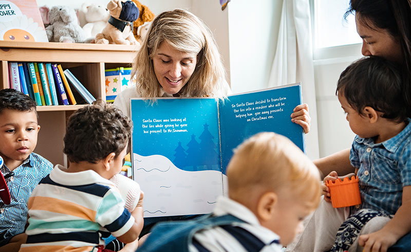 Nursery teacher reading a blue picture book to a group of toddlers during storytime