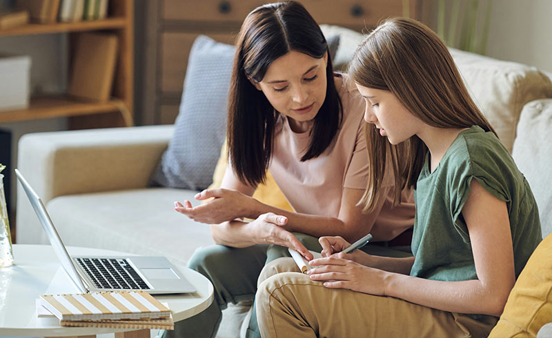 Woman helping a girl with a tablet for homework, laptop and notebook on a coffee table nearby