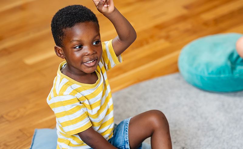 Smiling young boy sitting cross-legged on the floor, raising his hand during a group activity