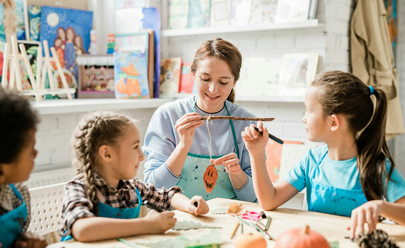 Craft instructor demonstrating a hanging autumn leaf craft to three children at a table with pumpkins and art supplies