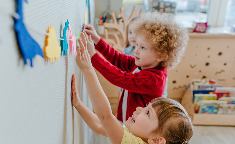 Preschool children decorating a classroom display with colourful paper figures