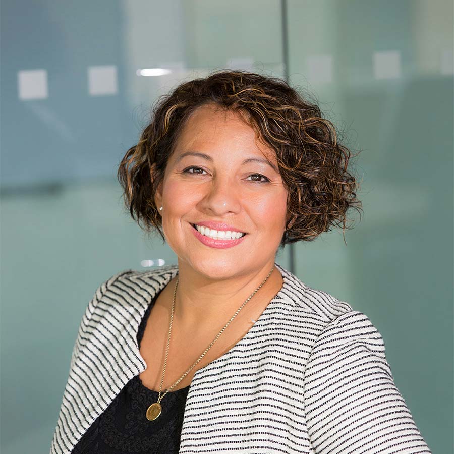 Smiling professional woman with short curly hair, striped blazer and gold pendant against a glass office background