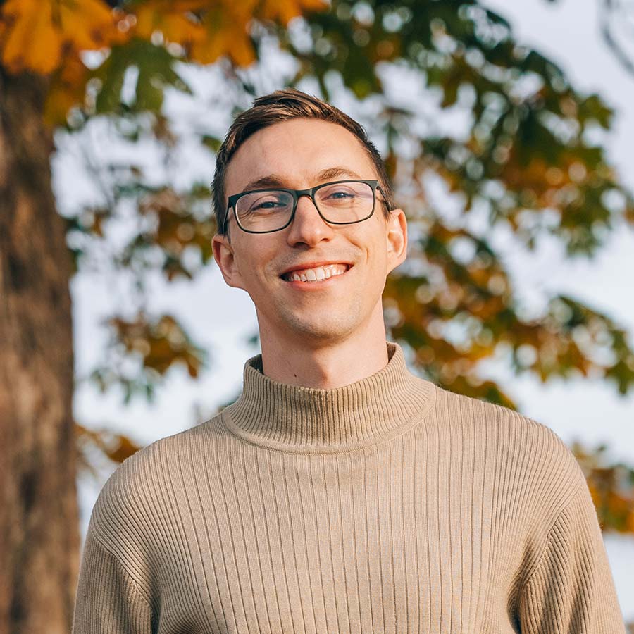 Smiling man with glasses wearing a beige ribbed roll-neck standing outdoors with autumn leaves behind him.