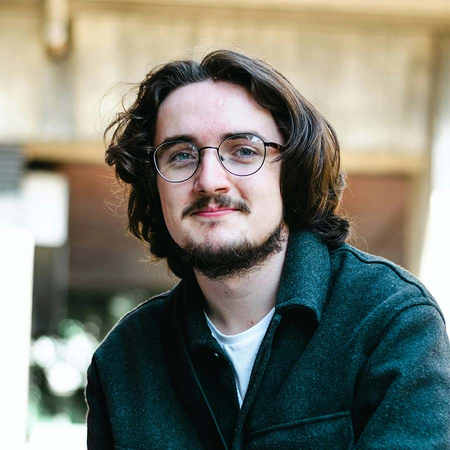 Young man with round glasses, shoulder-length hair and beard, wearing a dark green jacket and slight smile