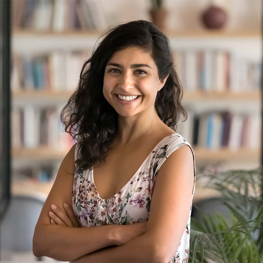 Smiling woman with arms crossed in front of a blurred bookshelf, wearing a floral top