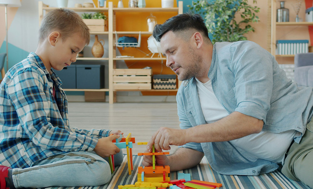 Father and young son building a colourful wooden block structure together on a striped rug in the living room