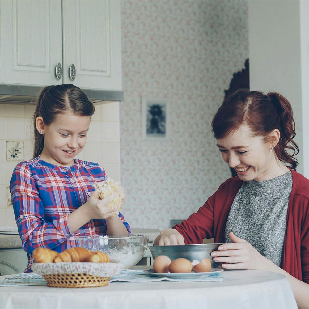 Woman and girl baking together, girl kneading dough at a kitchen table with mixing bowls, eggs and croissants