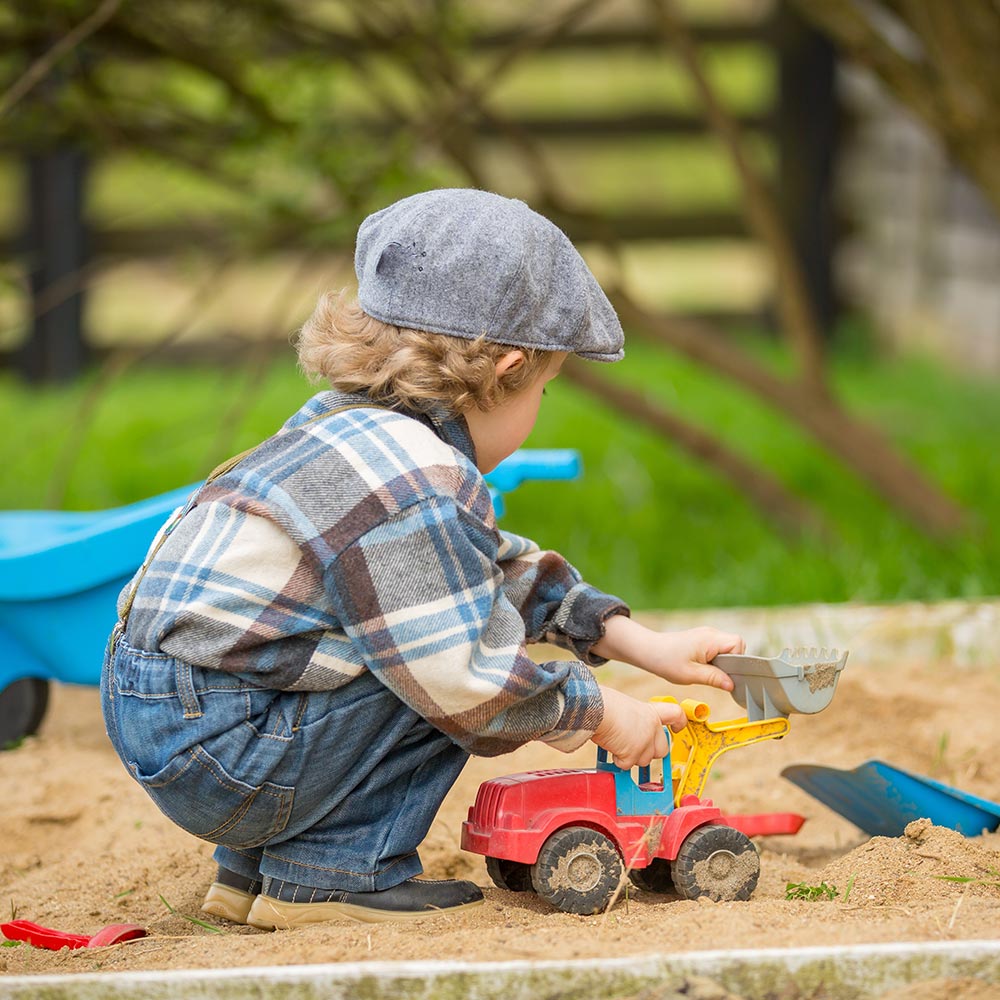 Child in a grey flat cap and plaid shirt crouching in a sandpit, pushing a red toy dumper truck
