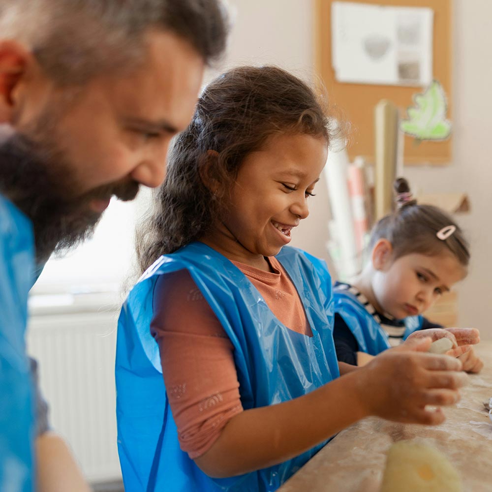 Girl smiling while modelling clay at a table, accompanied by an adult and another child in blue aprons.