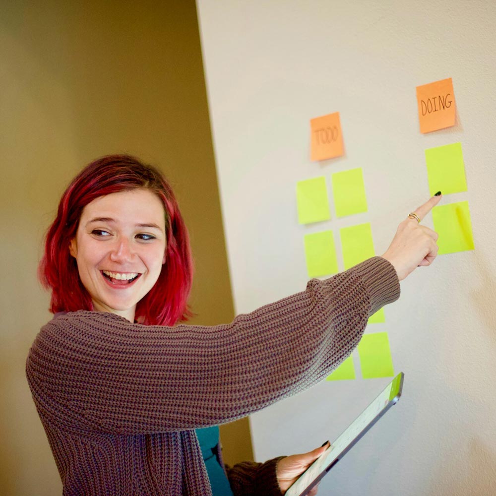 Smiling woman with pink hair pointing at Kanban-style sticky notes labelled "TODO" and "DOING" while holding a tablet