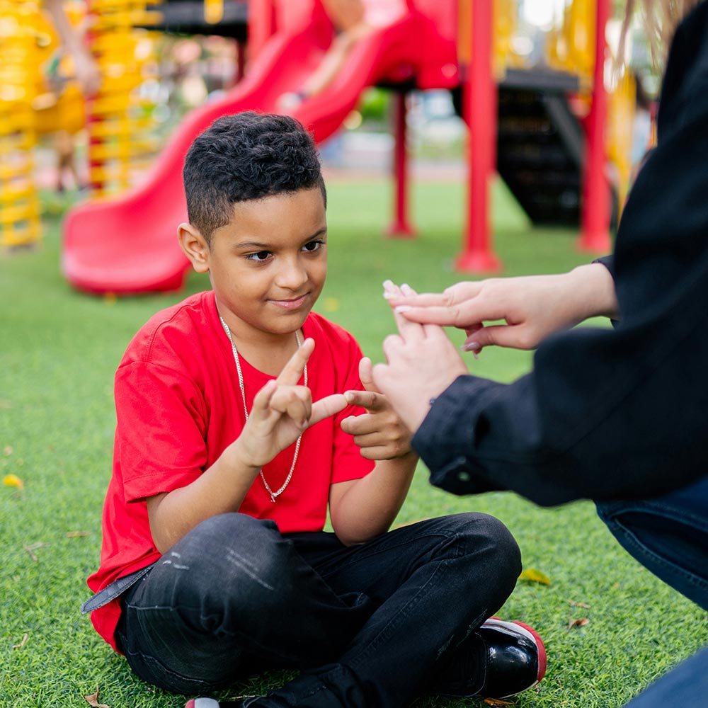 Boy practising sign language with an adult at a playground, seated on grass near a red slide