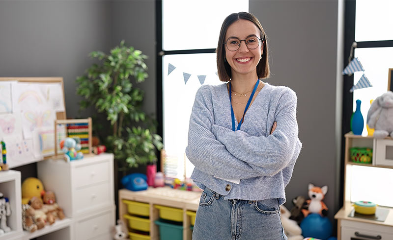 Smiling preschool teacher with glasses and lanyard in colourful classroom with toys