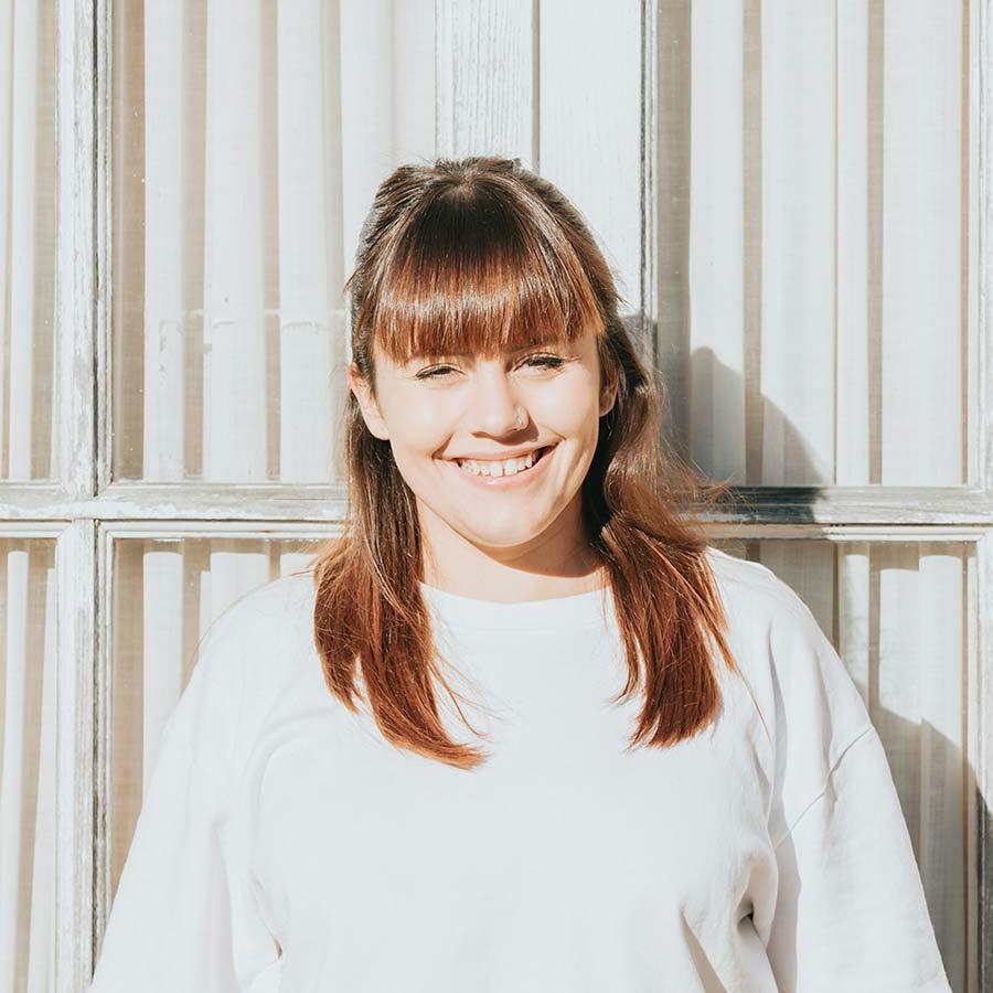Smiling woman with brown hair, fringe and nose stud, wearing a white top against a sunlit window