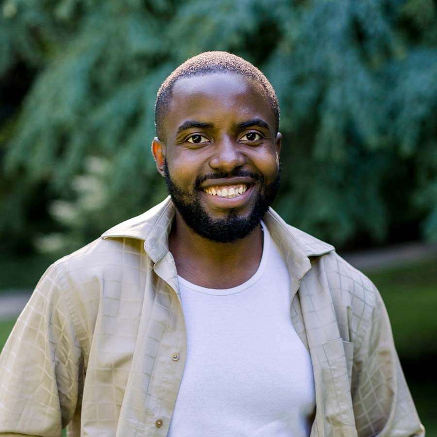 Smiling man wearing an open beige shirt over a white T‑shirt, standing outdoors with blurred trees behind him