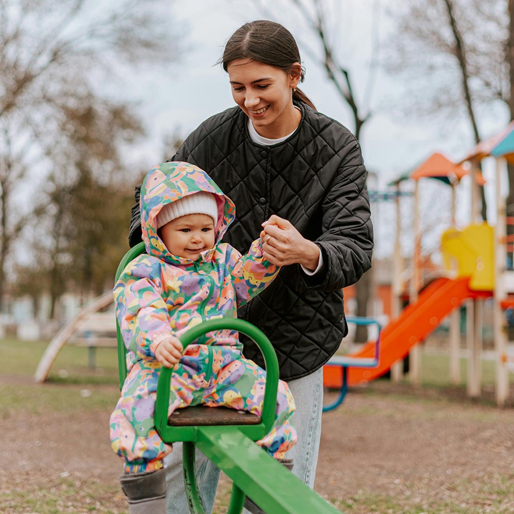Parent steadying toddler on a green seesaw at a playground, toddler wearing a colourful hooded snowsuit